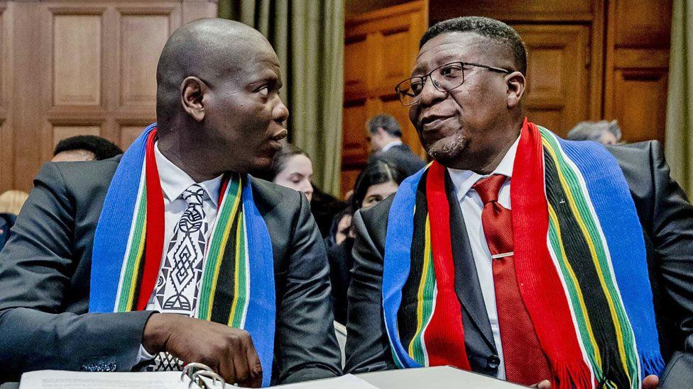 South African politician Ronald Lamola and another official - both in smart suits and with South African scarves around their necks - talk as they sit before a hearing at the International Court of Justice (ICJ) in The Hauge - January 2024