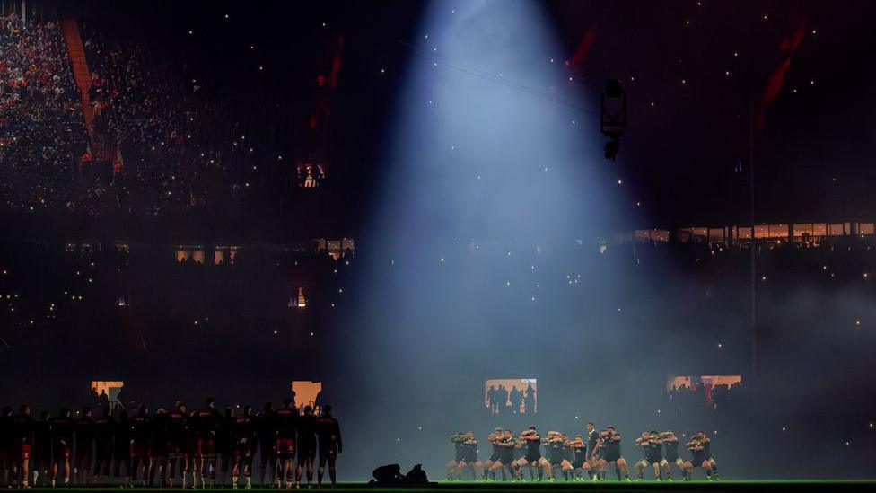 The All Blacks rugby team performing the haka under a dramatic spotlight in a dimly lit Principality Stadium in Cardiff.