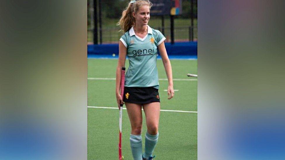 Clarissa Nicholls playing hockey at university. Miss Nicholls is wearing a light blue, short-sleeved, shirt bearing with the word "genei",  and long light blue socks. She is walking and carrying a pink, white and red hockey stick in her right hand.