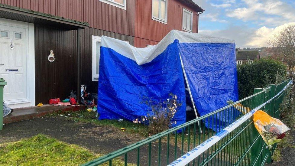 A forensic blue tent in a garden outside a property in Inverness. There is police tape and flowers hanging through green railings. The property is brown with a white door. There are items that look like children's toys scattered in the garden.