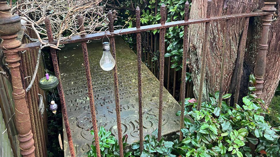 A flat headstone situated on the ground, with ornate engraving surrounded by rusted iron railings and plants