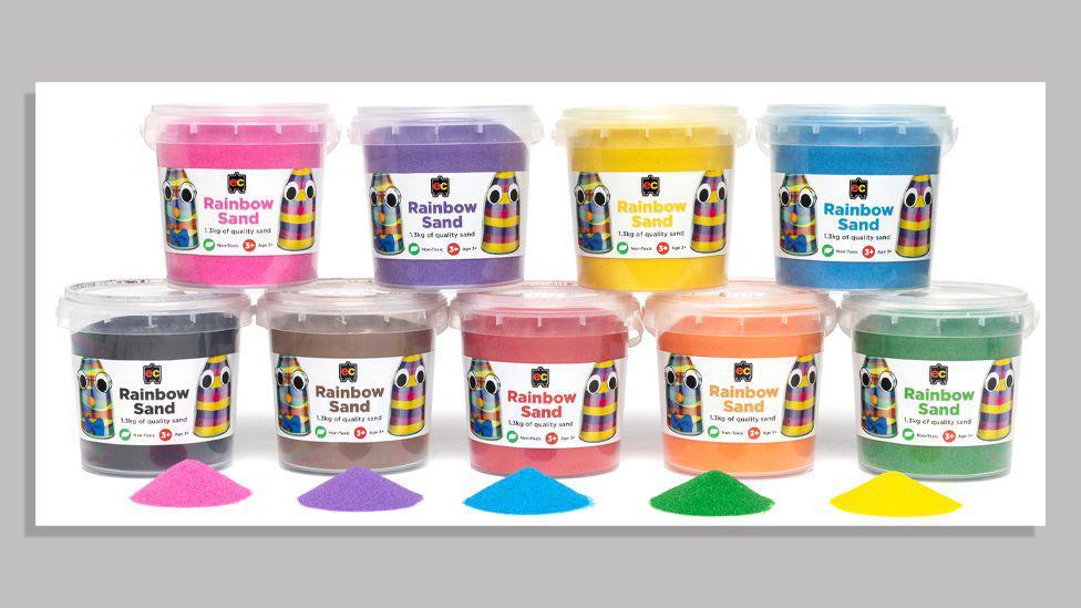 Nine different tubs of brightly coloured sand labelled as 'EC Rainbow Sand' are pictured against a white background. The colours are pink, purple, yellow, blue, black, chocolate brown, red, orange and dark green.