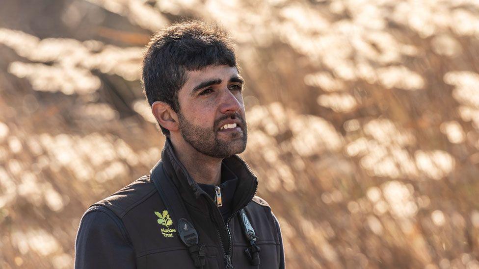 Ajay Tegala standing somewhat side on and looking towards the right. He has dark hair and a close cropped black beard. He is wearing a black gilet, with a yellow National Trust logo, over a black top and is standing in front of blurred out reeds.