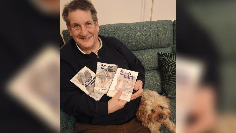 Michael Fleet - an older man with greying hair - smiles as he holds three of his Adventures of Bruin books in his arms as he sits on a green sofa next to his cavapoo, Maisie.