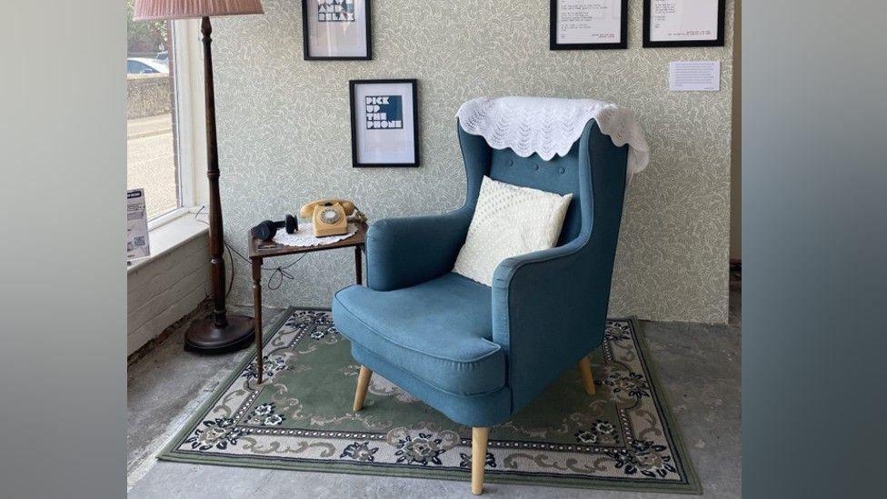 A blue chair in front of a wall. Next to it is a small table with a yellow rotary phone on it. There is a tall lamp on the ground next to the table.