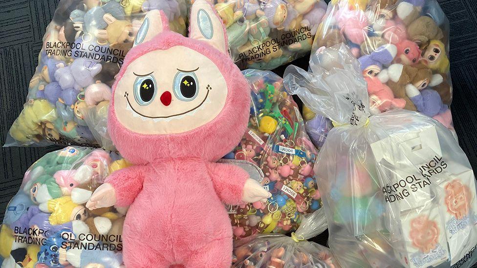 A large fake stuffed Labubu toy resting on top of bags of smaller dolls that have been seized. The large toy is pink with ears like a rabbit and a pale embroidered face with zigzag teeth, big eyes and a small red nose