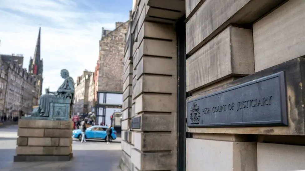 A shot of the entrance to the High Court in Edinburgh, a light-coloured building with an old statue in front of it. A sign reads "High Court of Justiciary"