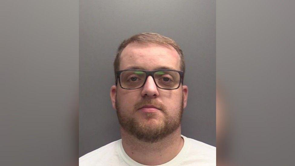 Darryl Sellars mugshot wearing black rectangular frame glasses with a beard and white t-shirt