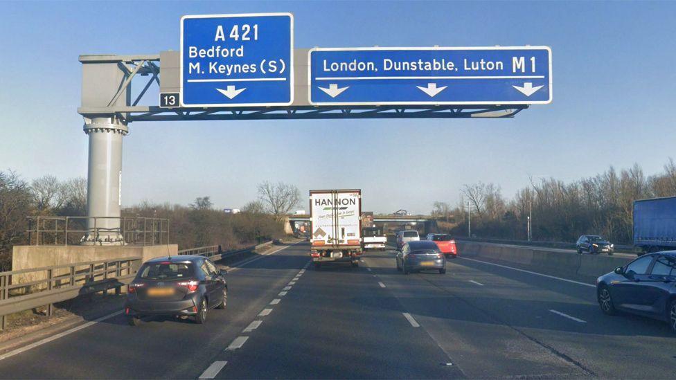 The exit for junction 13 of the M1 southbound, showing four lanes of traffic and a big sign above the road showing the right three lanes for the M1 - and the inside lane for the A421.