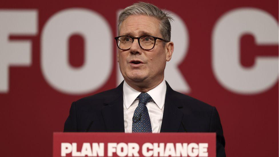 Sir Keir Starmer, with grey hair and wearing dark glasses, speaks at a podium in front of a red background 