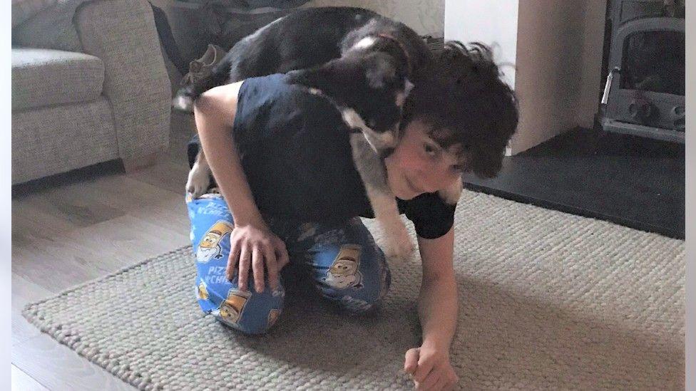 Josh is wearing a black t-shirt and blue pyjama bottoms. He is playing with his dog on a cream rug in the living room in front of an unlit stove fire. 