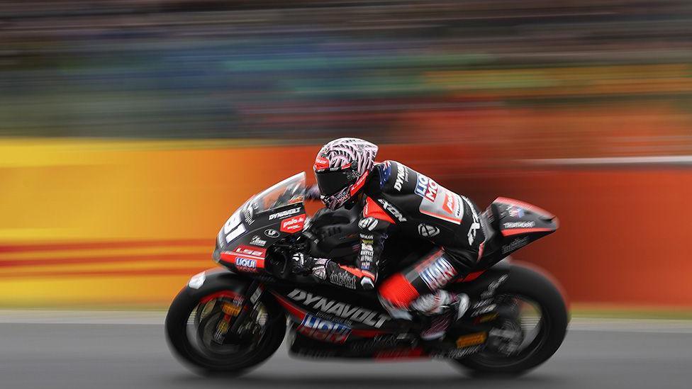 Motorcycle racer in black and red gear speeds along a track on a Dynavolt-branded bike, with motion blur emphasizing high speed against a colorful background.