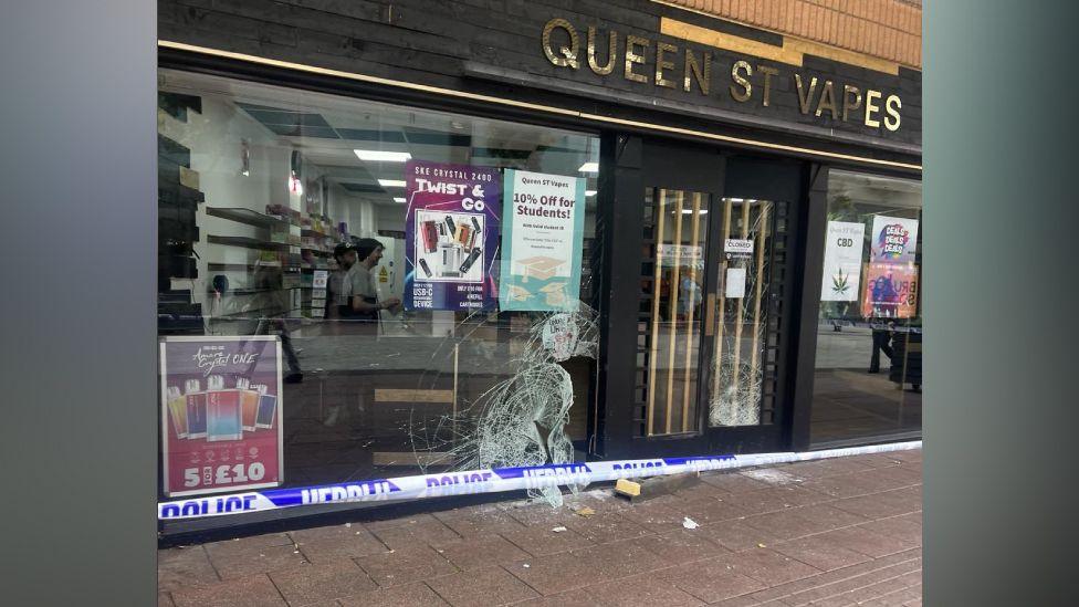 The outside of Queen Street Vapes shop, with a broken front window and police tape in front of it