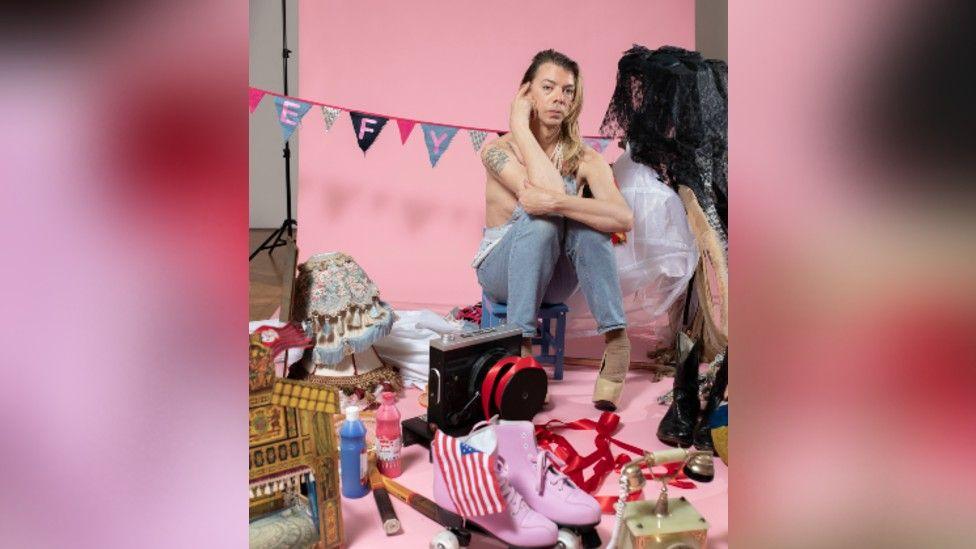 Tom Marshman wearing denim jean in a room with items symbolic of him. They include pink roller boots, a US flag, a lampshade, paints, net outfits and a boom box, all with a pink background.