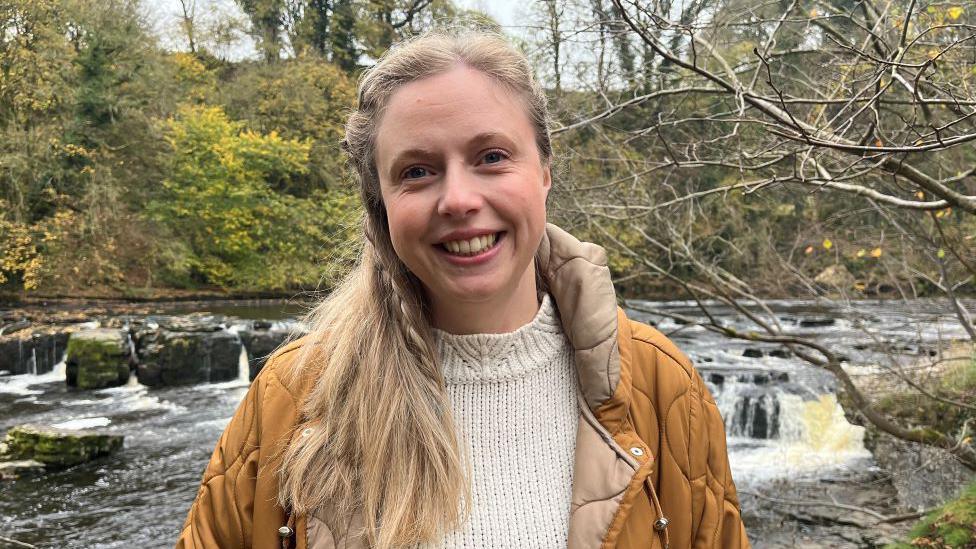 A woman with long blonde hair wearing a cream jumper and a mustard coat is standing in front of Aysgarth Falls.