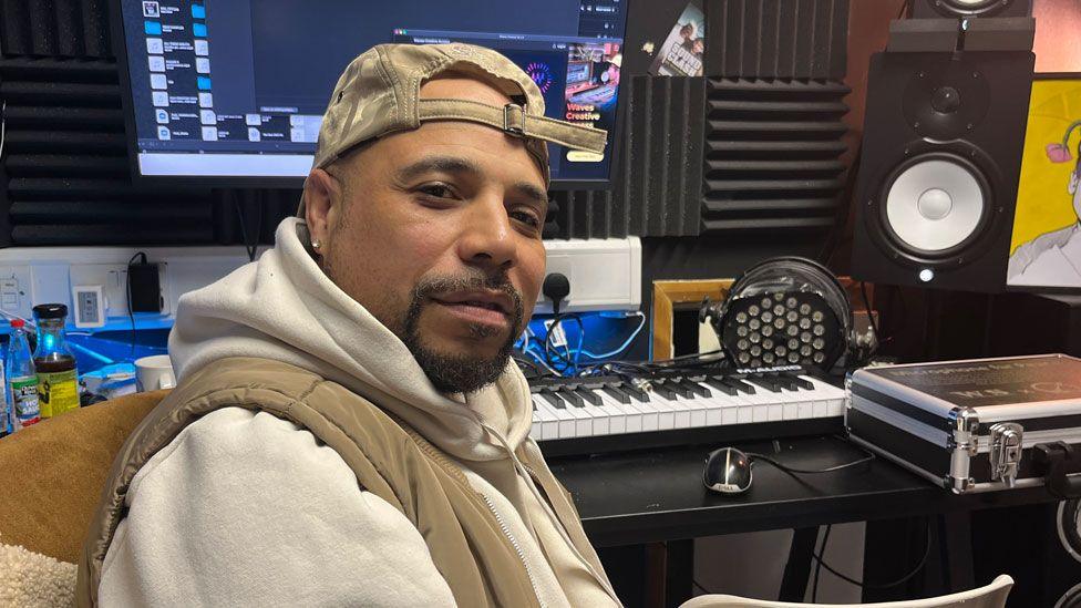 Karl Lewis - known as KD - sitting in a recording studio. He has turned sideways on and has a beige gillet over a cream hoodie, a neatly clipped black goatee beard and is wearing a beige baseball cap back to front. Behind him is a keyboard resting on a table, headphones and a computer screen.