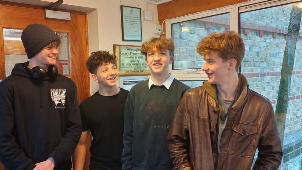 Four teenage boys in a row standing at the entrance to a school - they are in non uniform.