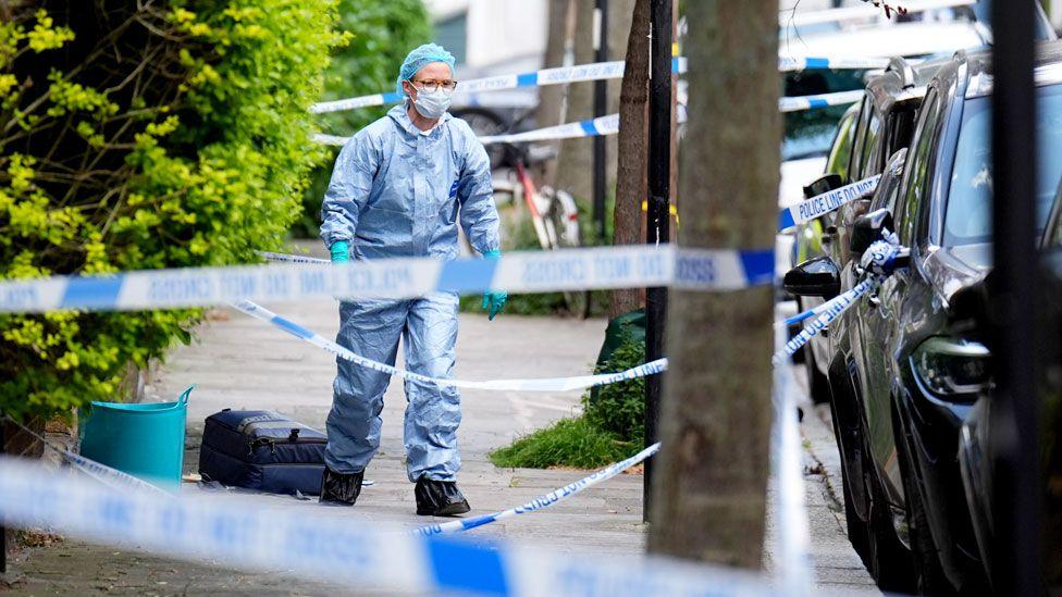 Police tape crosses a street between trees as a forensics worker walks down it