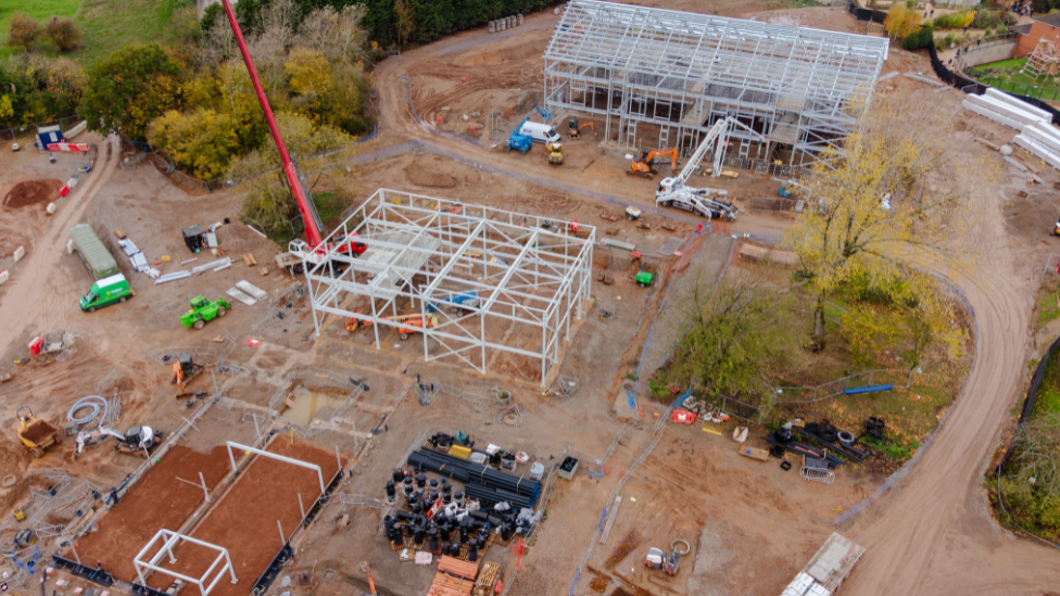 A birds eye image that shows the metal frames of two buildings on a building site. There are muddy roads and some trees.