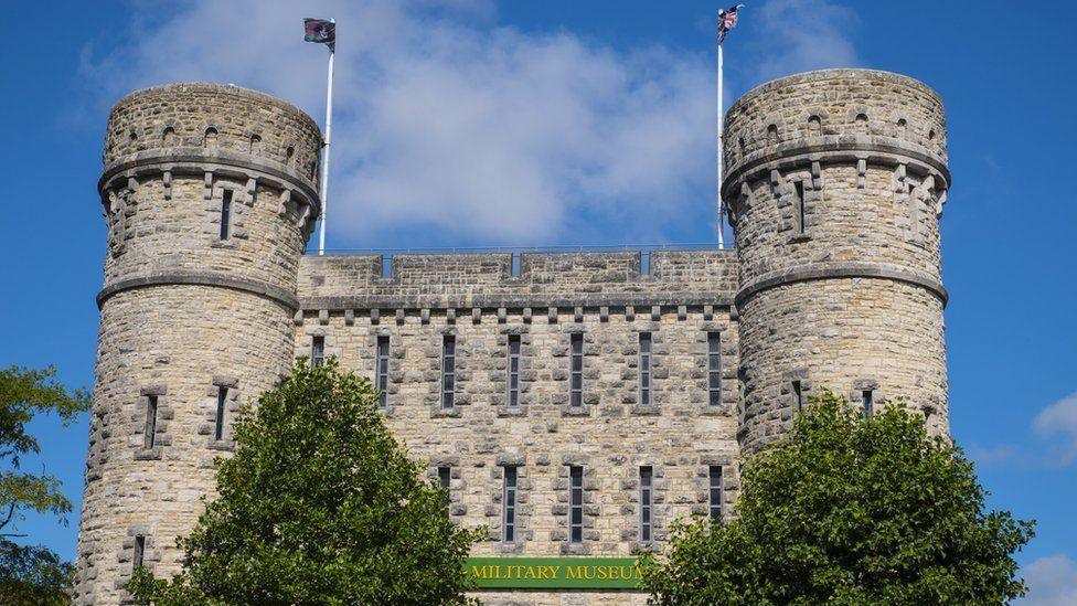 Stone castle-like building with flags flying ffrom two turrets, it has numerous thin slit-like windows and trees stand either side of its entrance.