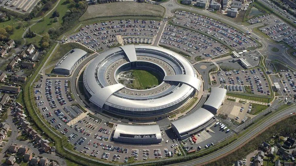 An aerial view of GCHQ headquarters, a circular, silver metal coloured building with several small units nearby, surrounded by car parks, a public road and a housing estate.