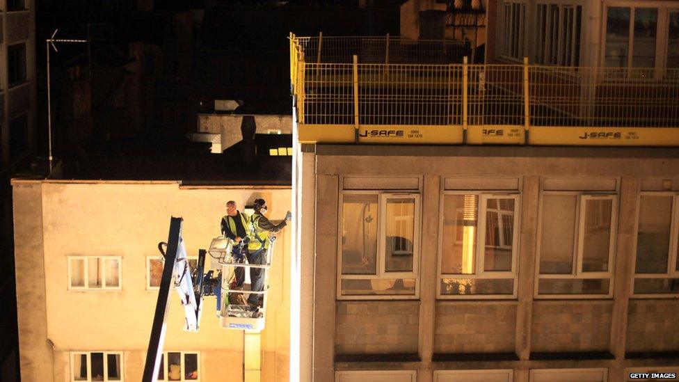 Two graffiti artists on a crane spray paint the top of a high rise building in Bristol.