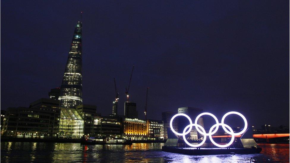 Olympic rings float past the Shard skyscraper with Waterloo Bridge in the background.