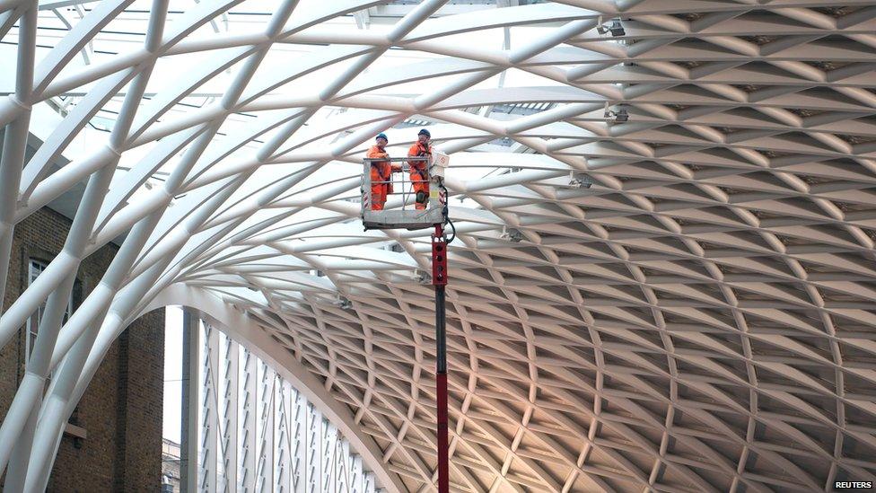A closer view of the glass roof over the tessellated white steel beams. Workers in orange overalls are on board a mechnical cherry-picker, inspecting the roof.