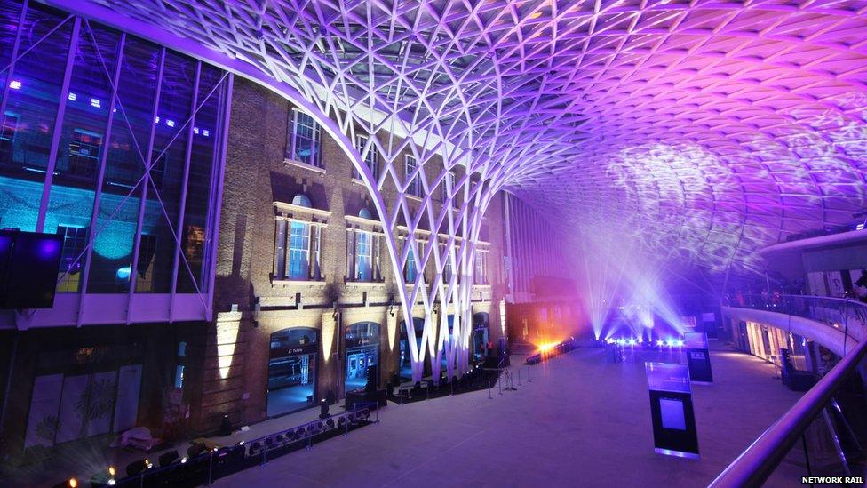 A view of the new King's Cross concourse. It is illuminated in vivid purple, blue and orange light. Beams of bright white light stretch through the vast concourse.
