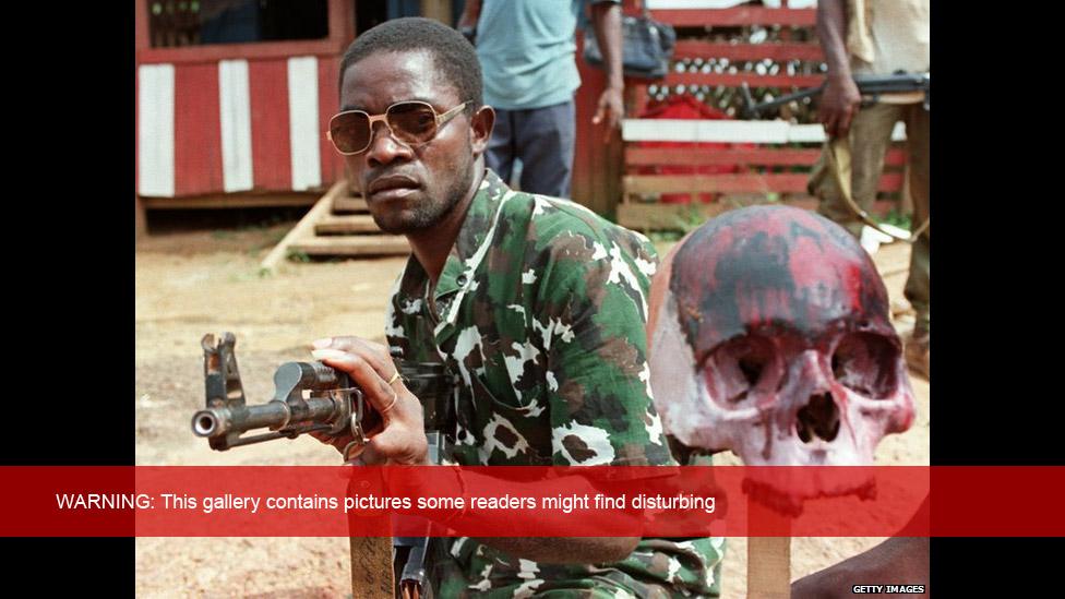 A rebel loyal to Charles Taylor holding Kalashnikov poses next to a painted skull of a Krahn ethnic soldier of president Samuel Doe, 15 May 1990 in Monrovia.