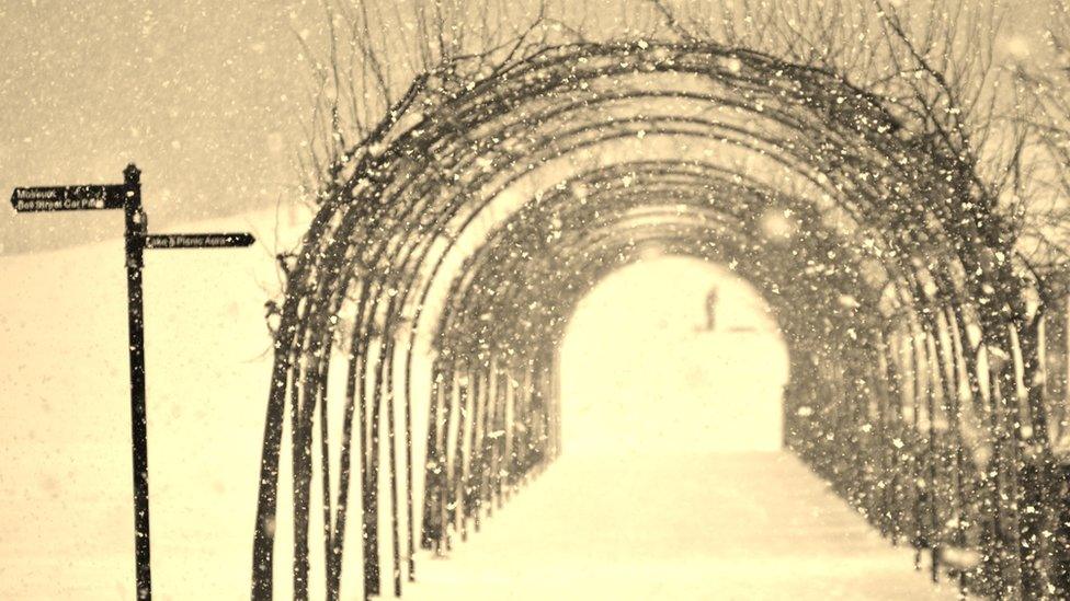 A sepia shot of an arch made out of branches with signposts to the left. All around is snow and it is snowing.