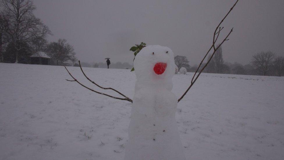 A small snowman with sticks as arms and a bright red nose is in the foreground. Behind is a very grey sky and a person walking with an umbrella.