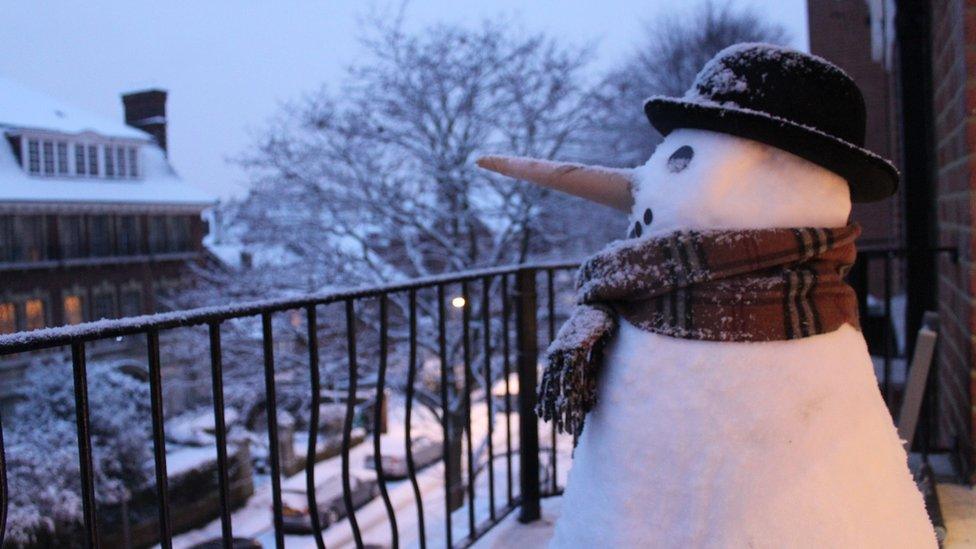 A snowman on a balcony looks out to the residential street. He is wearing a scarf and bowler hat and has a carrot for a nose.