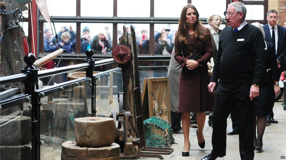 Duchess of Cambridge and John Vincent walking around National Fishing Heritage together with crowds peeking through the window in the background