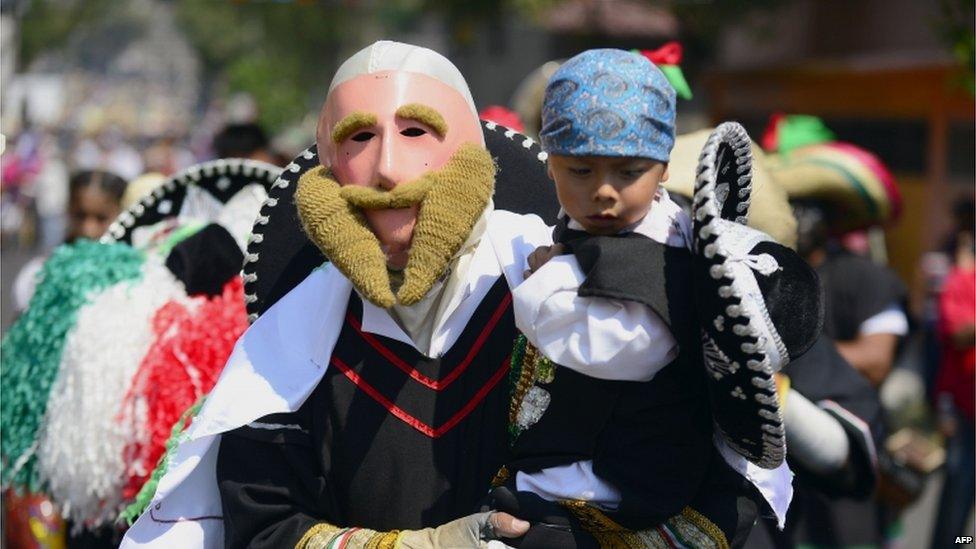 A man wearing a mask and costume is carrying a child at the Cinco de Mayo celebrations in Mexico City