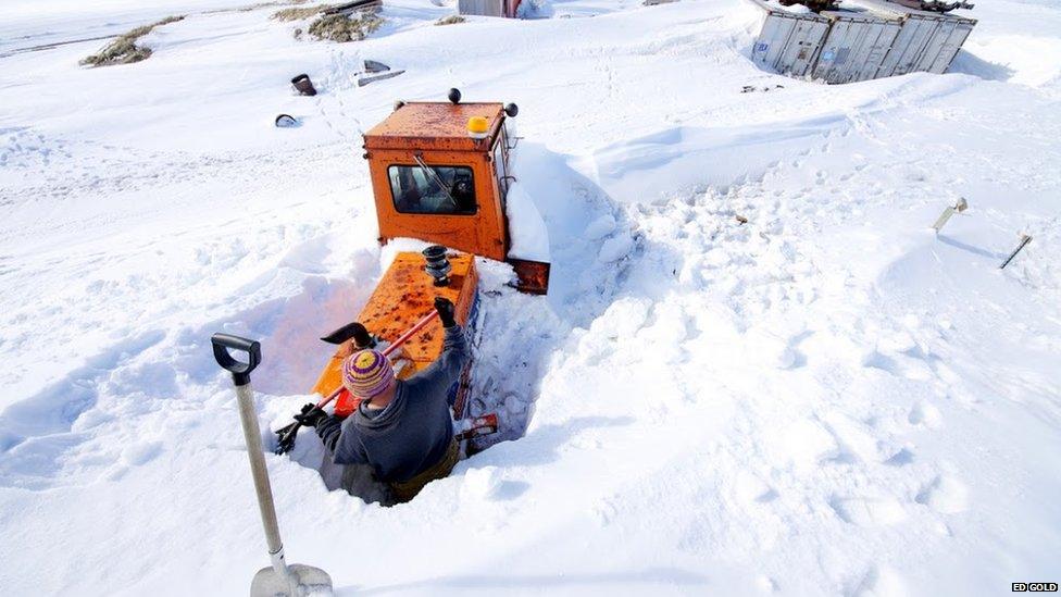 Sherman shovels snow away from a digger to get to a heater plug which will warm the engine overnight