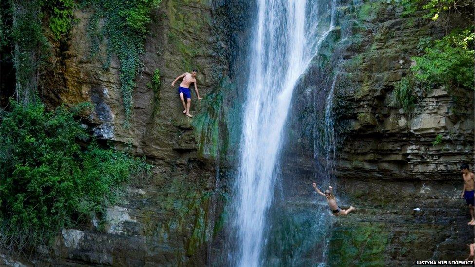 Boys in Georgia jumping off a rock face as water cascades down its surface