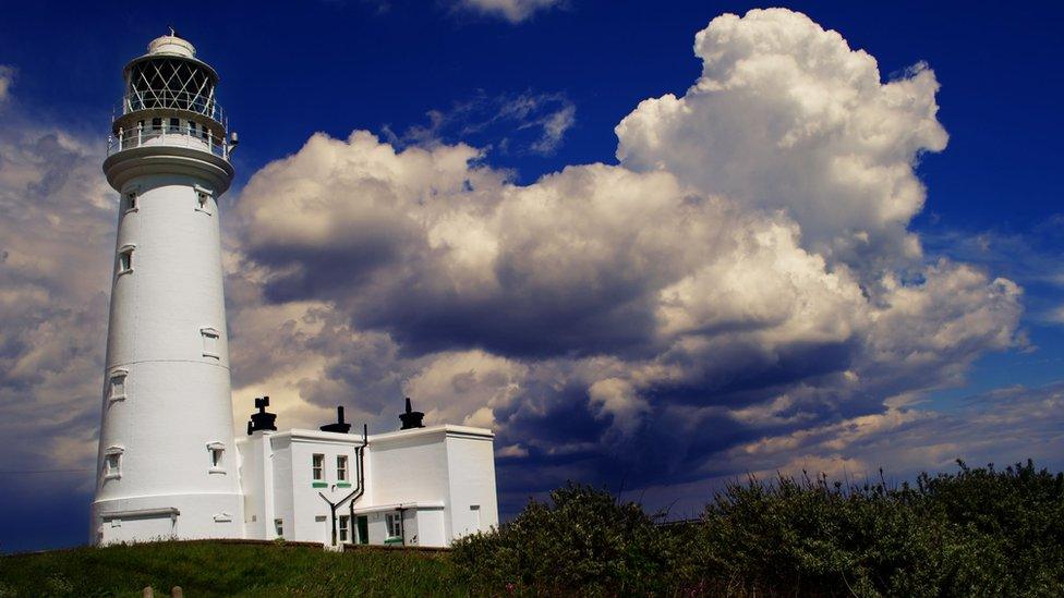 A white lighthouse and large white clouds in a blue sky above.