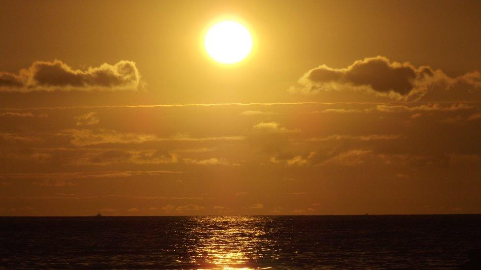 A yellow and gold sky, with a few clouds, holds a setting sun over the sea.