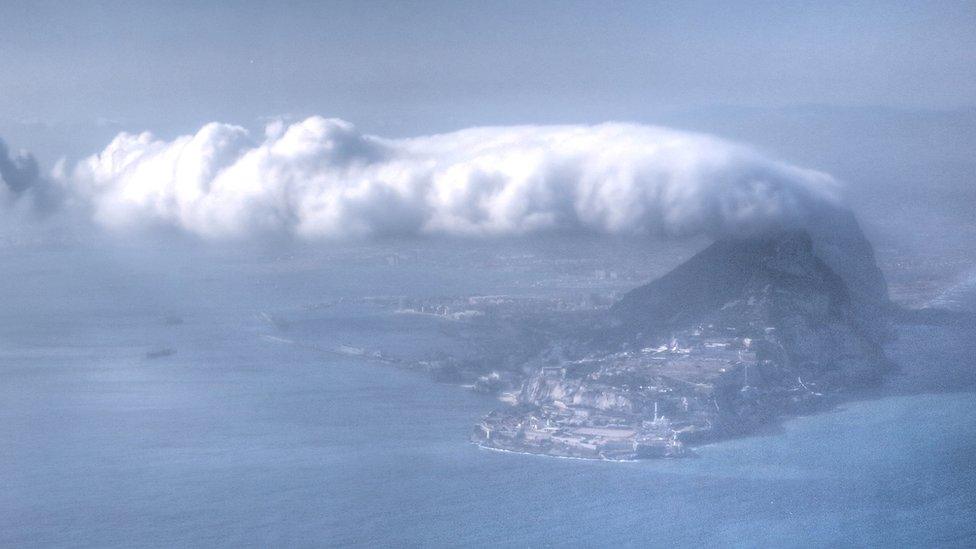 A photo from an aeroplane of a layer of thick cloud curving down over a rock peak on an island.