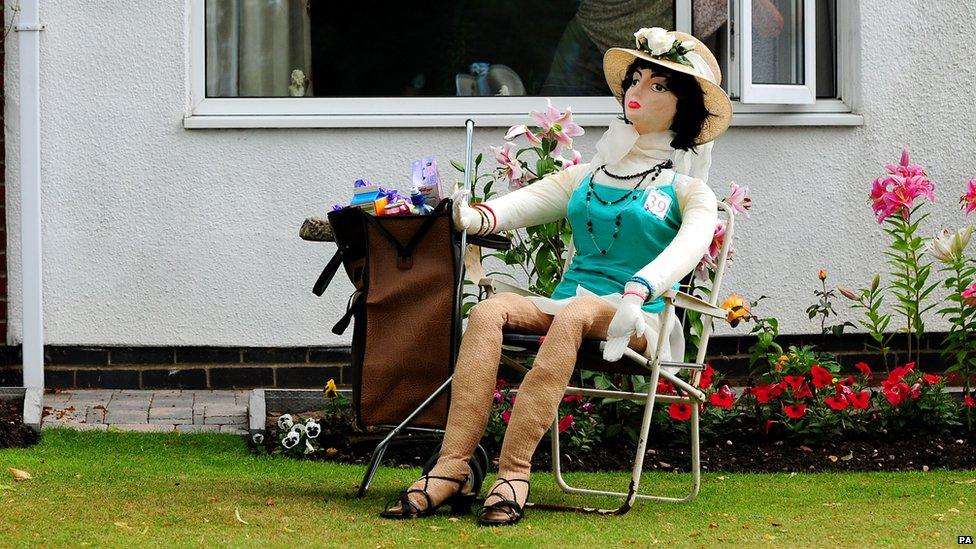 A scarecrow wearing a summer hat reclines in a garden chair next to her shopping trolley.