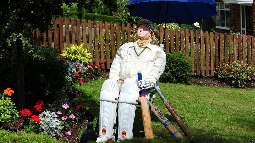 A scarecrow dressed as a cricket player sits underneath and umbrella with his bat.