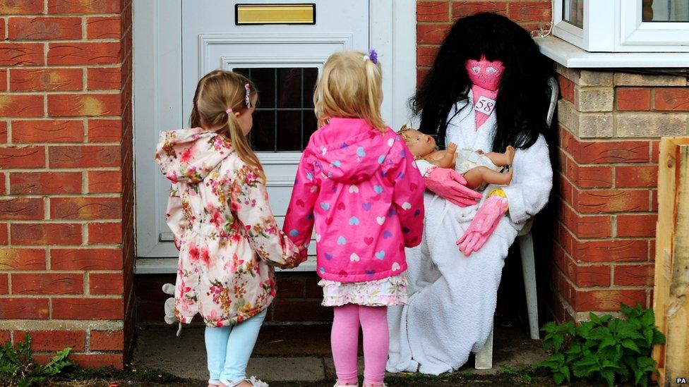 Children look at a pink scarecrow dressed as Kate Middleton with a brown wig. The scarecrow is cradling a baby.