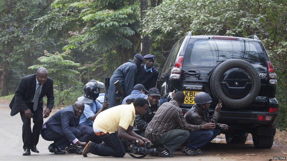 Police officers and members of the media take cover at a distance from the Westgate Shopping Centre after continuous gunfire was heard on September 23, 2013.