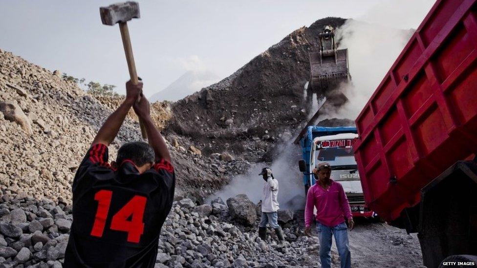 A worker using a hammer to break the rock as excavator fills a truck with sand in Gendol River riverbed on 6 September 2013 in Yogyakarta, Indonesia