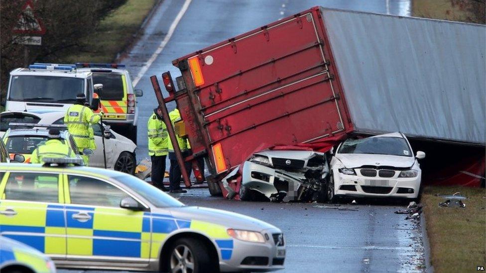 An overturned lorry lying on top of two damaged cars on an A-road