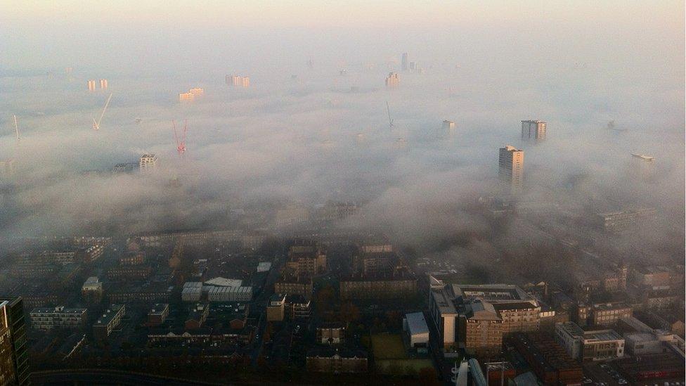 Mist dominates a city panorama. Tall flats and buildings poke out above the blanket.