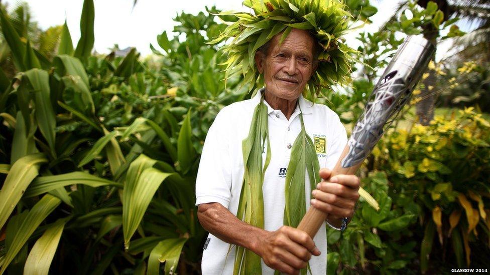 An elder in a traditional floral headdress holds the Queen's Baton in Rarotonga, Cook Islands.