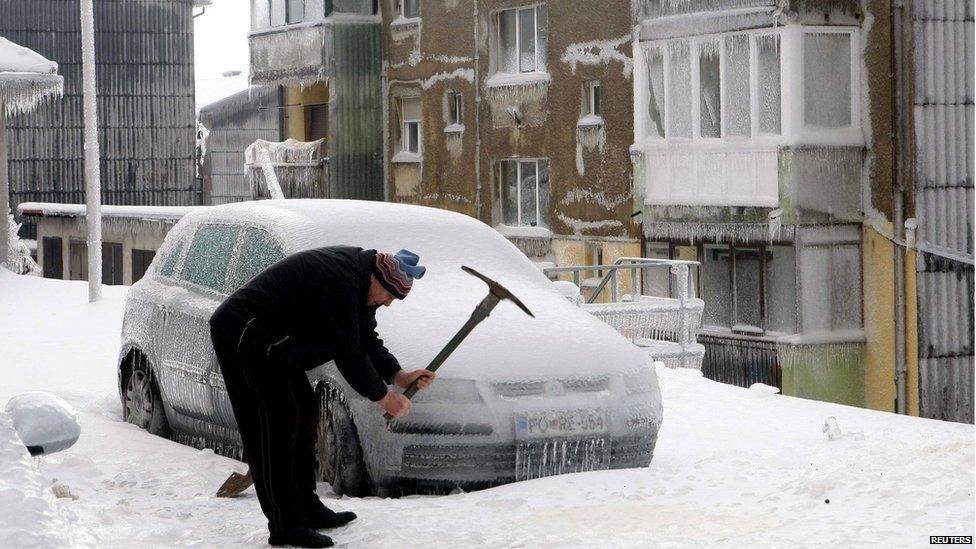 A man smashes ice with a pickaxe next to car covered with ice