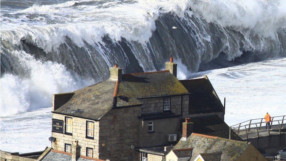 A huge wave heads towards shore, near a pub and houses. A man stands on a bridge-like structure looking at the wave coming in.